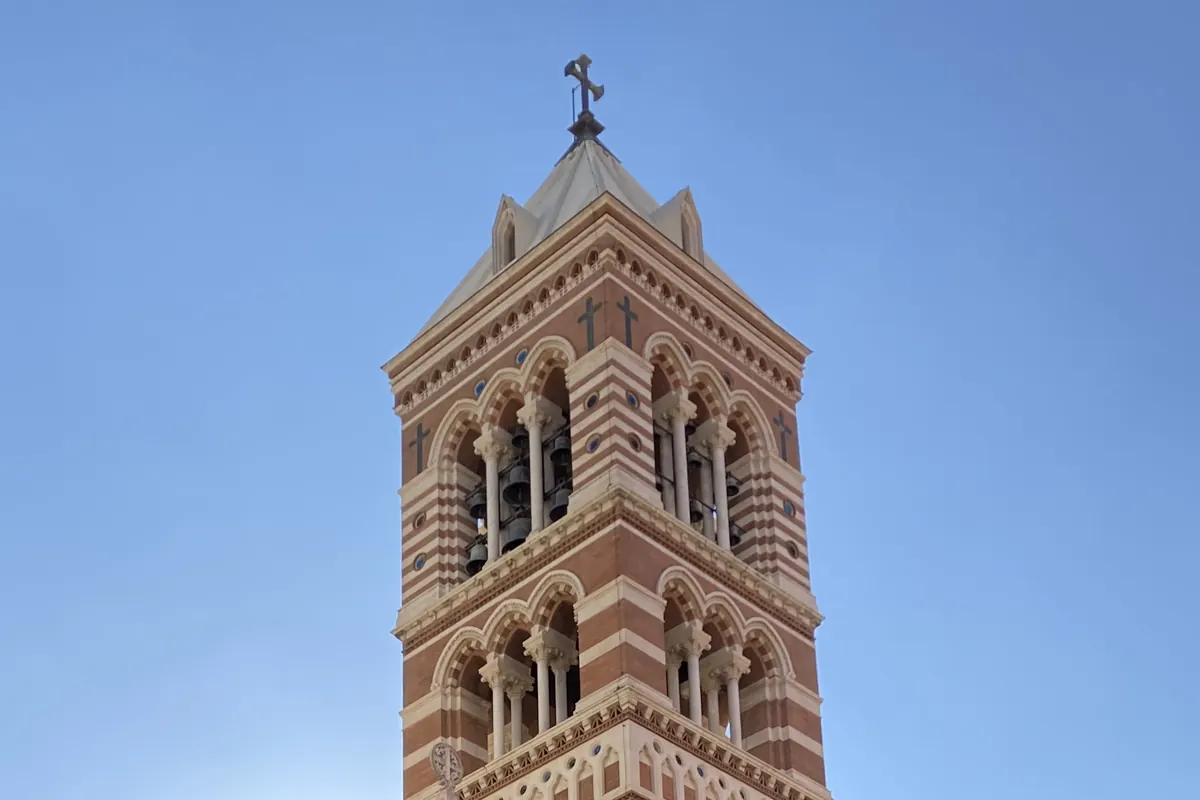 The striped brick and stone bell tower of St Paul's Within the Walls against a clear blue sky, with a cross on its pointed roof.
