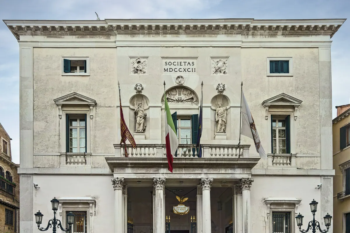 The pale stone front of the Teatro La Fenice in Venice with a columned entrance, a balcony and three flags.
