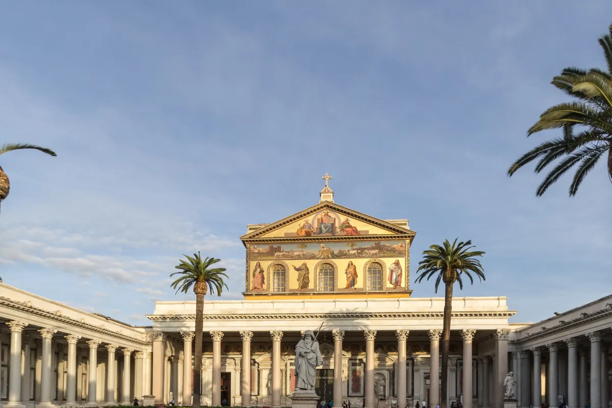 A courtyard of white columns and palm trees leading to a basilica front topped by a gold mosaic gable.